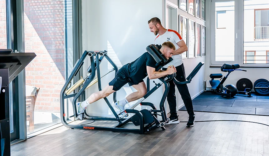 Der Sportwissenschaftlicher Patrick Reich trainiert einen Kunden in einem Sportgerät für Kraft-Ausdauertraining.