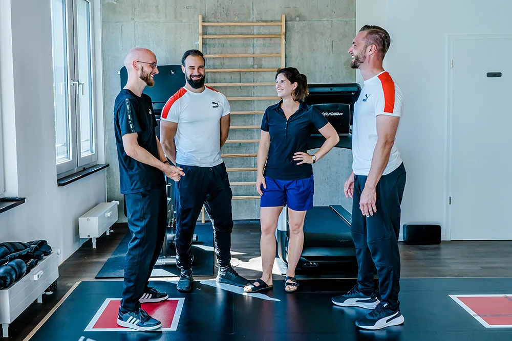 Ein Gruppenfoto von vier Physiotherapeuten der Physiotherapie ALC-Physiolab. Die Gruppe steht im Halbkreis und die Physiotherapeuten und Physiotherapeutinnen unterhalten sich.